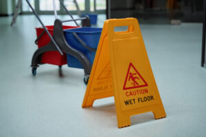 A yellow "Caution Wet Floor" sign stands on a light-colored floor next to a mop bucket with two sections, one red and one blue. The bucket is mounted on a wheeled frame. The clear display highlights proactive measures to prevent slips and trips, emphasizing the importance of workplace safety.
