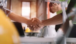 Two people shaking hands in a close-up view, suggesting a successful agreement or partnership. A construction site setting is implied by the presence of a white hard hat, safety vest, and construction plans on a table in the background, emphasizing the importance of safety and fall prevention.