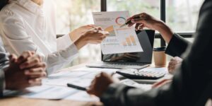 Two people are sitting at a table, reviewing a business report with charts and graphs. One person is holding the report while the other is pointing to it with a pen, discussing insights. A laptop, calculator, and takeaway coffee cup are on the table. Natural light is coming from a window.