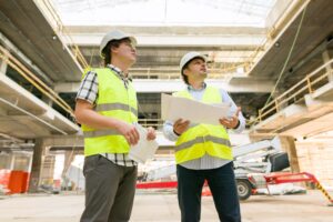 Two construction workers in safety vests and hard hats are observing something in a large, partially constructed building. They are holding blueprints and appear to be discussing the project while standing amid scaffolding and equipment, ensuring height safety protocols are followed around the site.