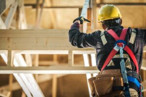 A construction worker wearing a hard hat, safety harness, and tool belt is climbing a wooden structure inside a building under construction. The worker is viewed from behind, highlighting the safety equipment and personal fall arrest systems in place.
