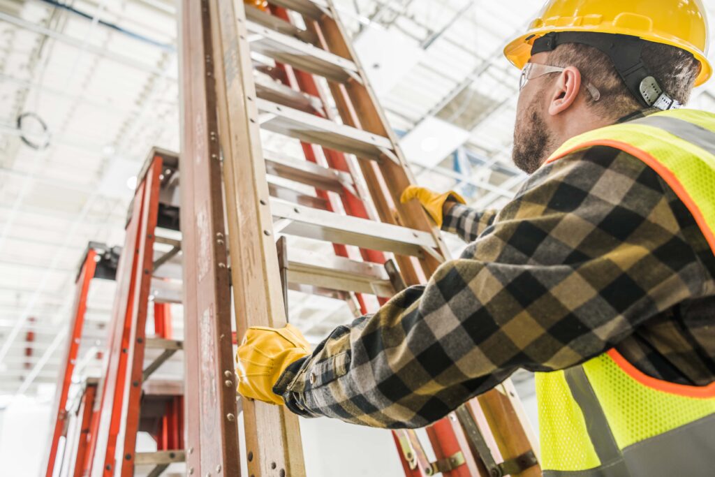 A construction worker wearing a yellow hard hat, safety vest, gloves, and a plaid shirt is holding a brown ladder. The background includes other ladders and a high ceiling with lighting fixtures, indicating an indoor work site focused on hazard detection.