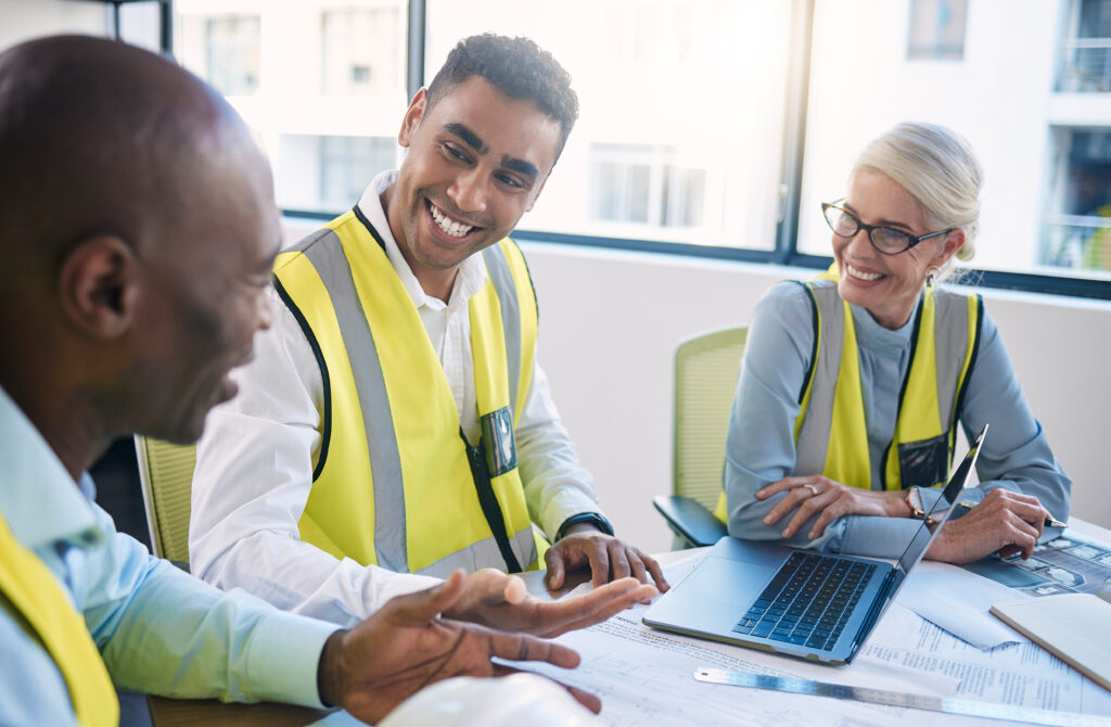 Three construction workers wearing yellow safety vests sit around a table in an office, smiling and engaged in conversation. A laptop is open on the table along with blueprints and documents, including workplace certifications. Large windows in the background let in ample natural light.