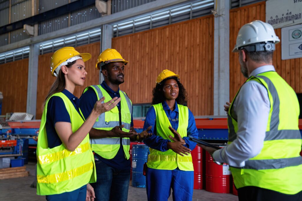 Four people wearing safety vests and hard hats are conversing inside an industrial warehouse. Three workers are engaged in discussion with a supervisor, who is holding a tablet. Emphasizing workplace safety, the background features storage containers and industrial equipment.