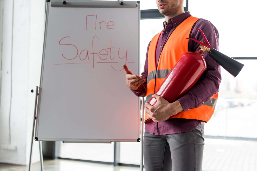 A person wearing an orange safety vest stands in front of a whiteboard that has "Fire Safety" written on it in red. They are holding a red fire extinguisher in one arm while gesturing with their other hand, emphasizing the importance of workplace emergency training during an indoor session.