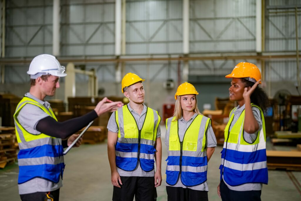 Four workers in reflective vests and hard hats are standing in a warehouse. One worker, holding a clipboard, appears to be giving instructions on advanced techniques while the others listen attentively. The background shows warehouse shelves and industrial equipment, highlighting their commitment to occupational safety training.