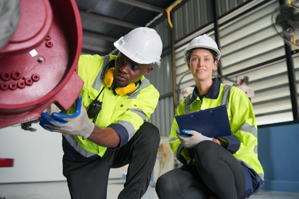 Two workers in high visibility jackets and white safety helmets are inspecting machinery indoors. One, a man, is adjusting a red machine part. The other, a woman, is holding a blue clipboard and smiling. Both are focused on hazard detection and working in a well-lit industrial environment.