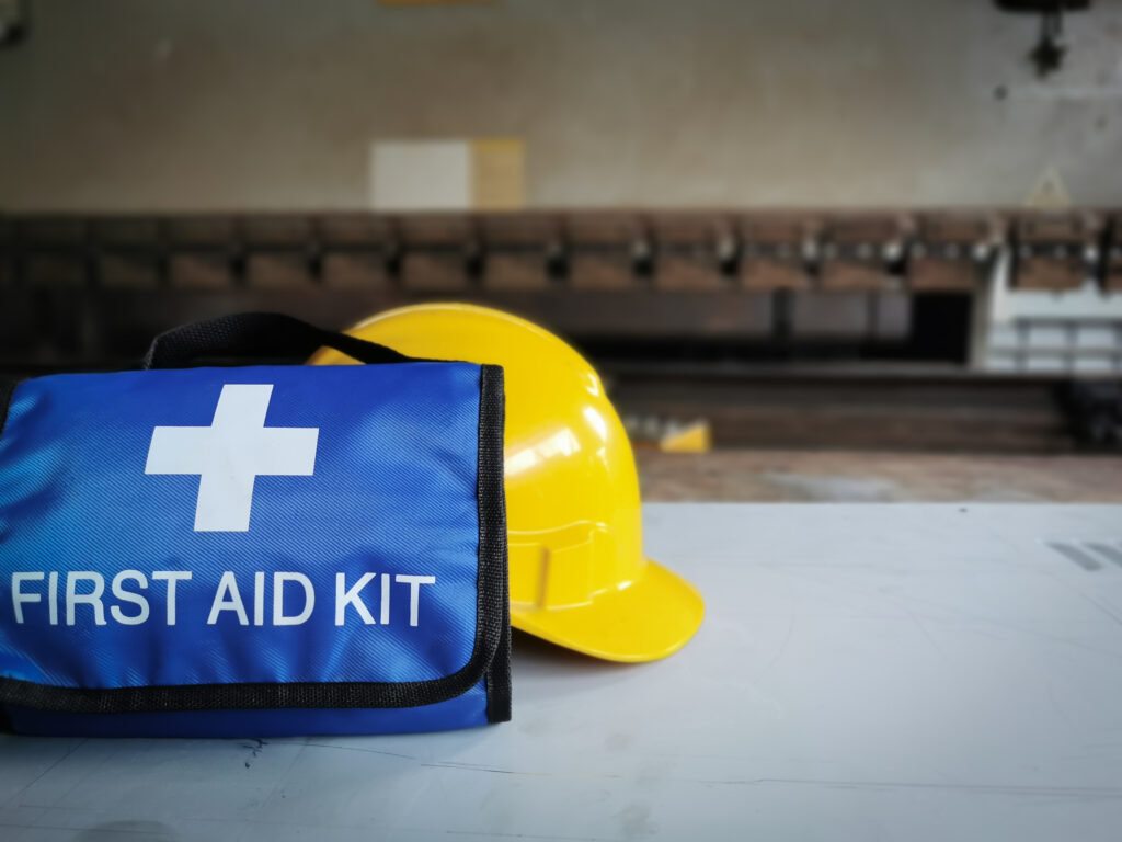 A blue first aid kit with a white cross symbol and text "FIRST AID KIT" is placed on a table next to a yellow hard hat, emphasizing the importance of workplace safety. The background shows industrial equipment and a workshop environment, highlighting the necessity of accessible medical supplies.