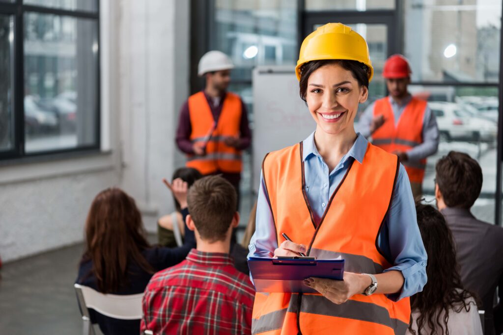 A woman in a hard hat and orange safety vest stands smiling with a clipboard in hand. She is in a room with several other people, some also wearing safety gear, sitting and listening to a presentation on hazard awareness.