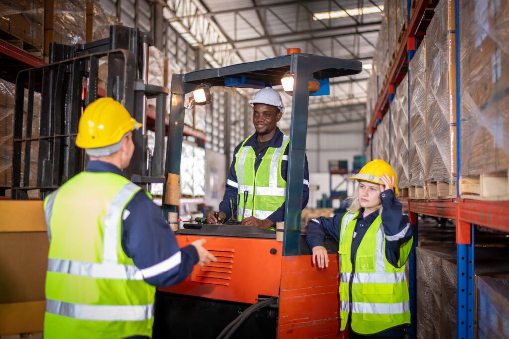 In a well-lit warehouse, a worker with effective forklift training operates a forklift while two other workers, all wearing reflective safety vests and hard hats, stand nearby. The shelves are filled with large boxes, showcasing the importance of safety in such environments.