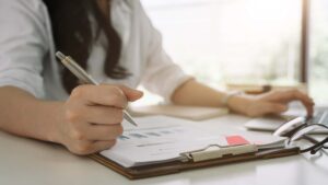 A person is seated at a desk, holding a pen and writing on a document attached to a clipboard. Their other hand is typing on a laptop. The scene includes a pair of glasses and some charts displaying key statistics on the desk, with soft natural light illuminating the workspace.