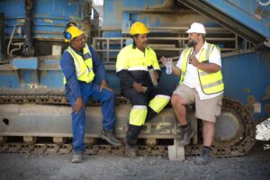 Three construction workers on a break, sitting in front of heavy machinery. Two are wearing blue and neon yellow outfits with helmets, while the third in khaki shorts, a white shirt, and a white helmet gestures while talking. All wear safety vests, ensuring hydration by sipping water to maintain health.