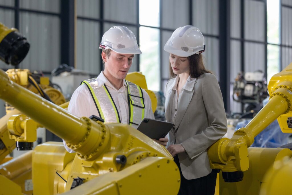 Two people in white hard hats are examining industrial machinery in a factory setting, conducting Machinery Assessments. One holds a clipboard while the other looks on. They are surrounded by yellow robotic arms and various other equipment. Both people wear safety vests and business attire, emphasizing Workplace Safety.