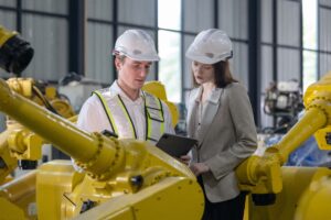 Two people in white hard hats are examining industrial machinery in a factory setting, conducting Machinery Assessments. One holds a clipboard while the other looks on. They are surrounded by yellow robotic arms and various other equipment. Both people wear safety vests and business attire, emphasizing Workplace Safety.