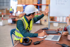 A construction worker wearing a white hard hat and reflective vest sits at a table with a laptop, notebook, and radio. He points towards a whiteboard, discussing Virtual Workplace Safety Training with colleagues in a warehouse setting. Another person is visible, raising their hand.