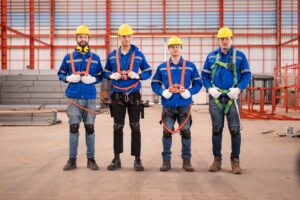 Four construction workers stand in a warehouse, facing the camera. They wear blue uniforms, yellow hard hats, safety glasses, gloves, and knee pads. Each worker is equipped with a safety harness for harness protection. Stacks of materials and red steel structures are seen in the background.