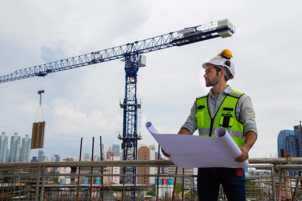 A construction worker in a neon yellow safety vest and white helmet holds blueprints while standing at a construction site. A large crane is in the background, with buildings and a cloudy sky visible. The worker, adhering to best practices for outdoor workers in hot climates, appears to be inspecting the site.