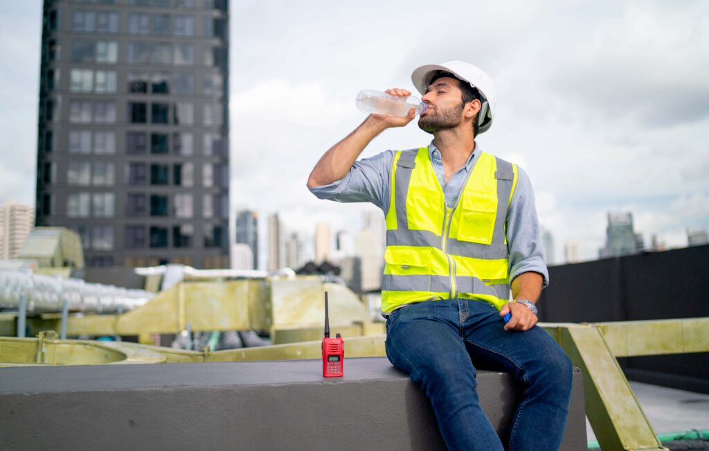 A construction worker wearing a white helmet, yellow safety vest, blue shirt, and jeans sits on a concrete ledge. He is drinking water from a bottle, with a walkie-talkie placed beside him. Emphasizing OSHA Heat Standards in hot environments, the cityscape with high-rise buildings is visible in the background.