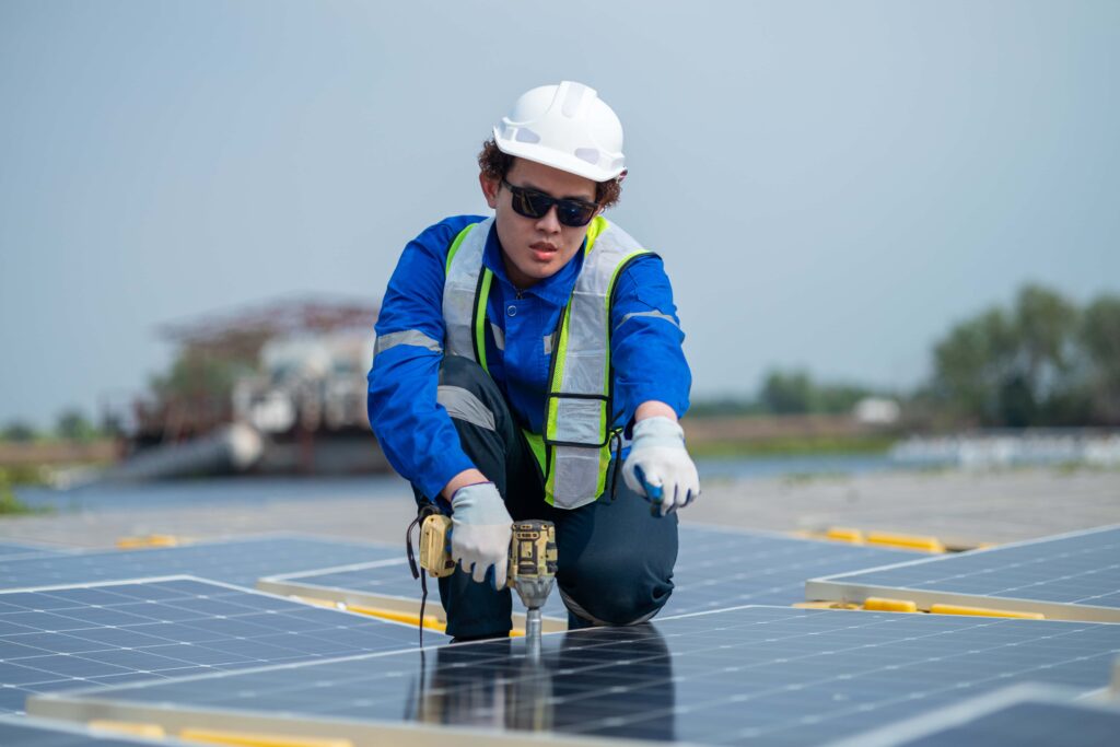 A worker wearing a white helmet, sunglasses, blue uniform, and safety vest kneels on a rooftop, using a power drill to install solar panels. With an emphasis on workplace safety, trees and industrial structures are visible in the background.