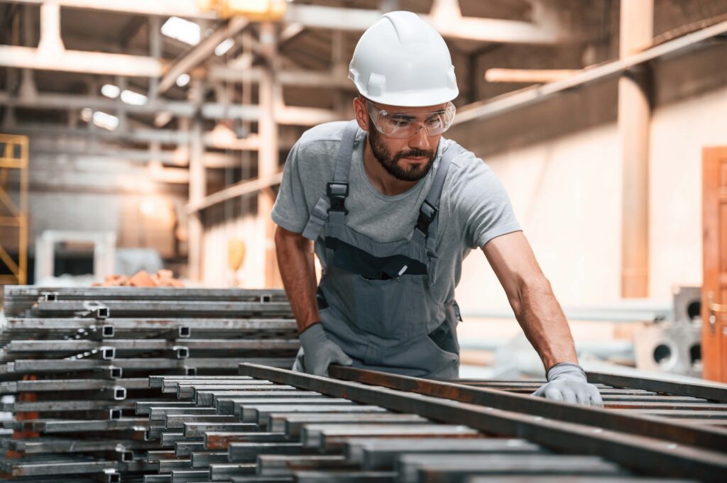 A man wearing a white hard hat, safety glasses, and grey overalls is working in a factory. He is handling metal beams, which are stacked in front of him. The background shows an indoor industrial setting with various equipment and structures; heat safety precautions are clearly observed.