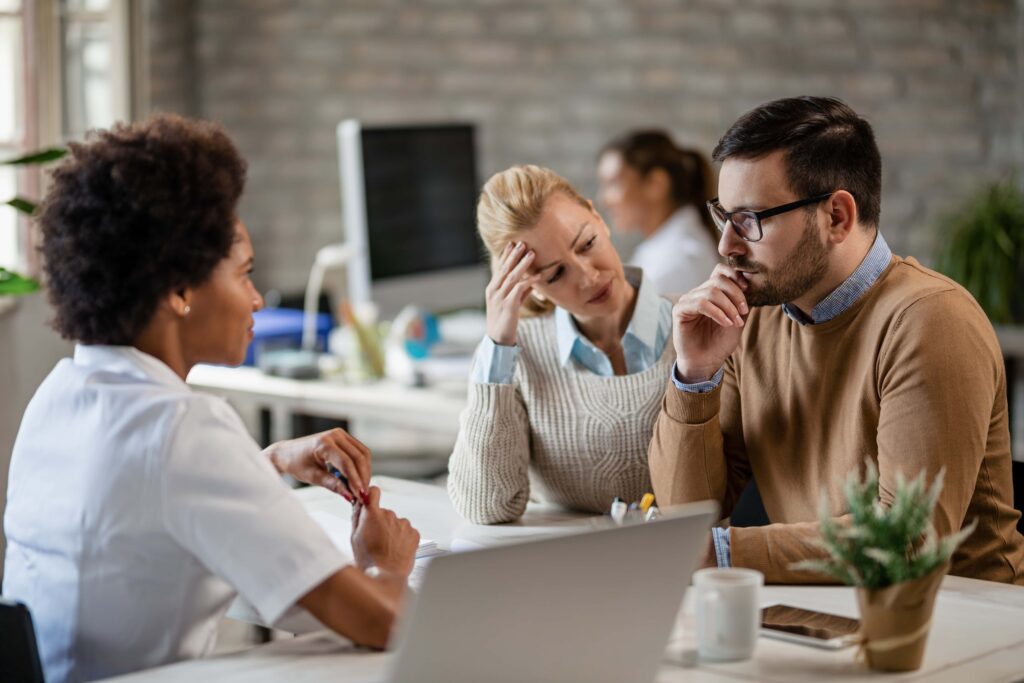 Three people are having a serious discussion in an office. A woman with curly hair in a white lab coat sits across from a man in glasses and a woman with blonde hair, who both look concerned. Stress levels seem high as they discuss mental health. A laptop and a potted plant are visible on the desk.