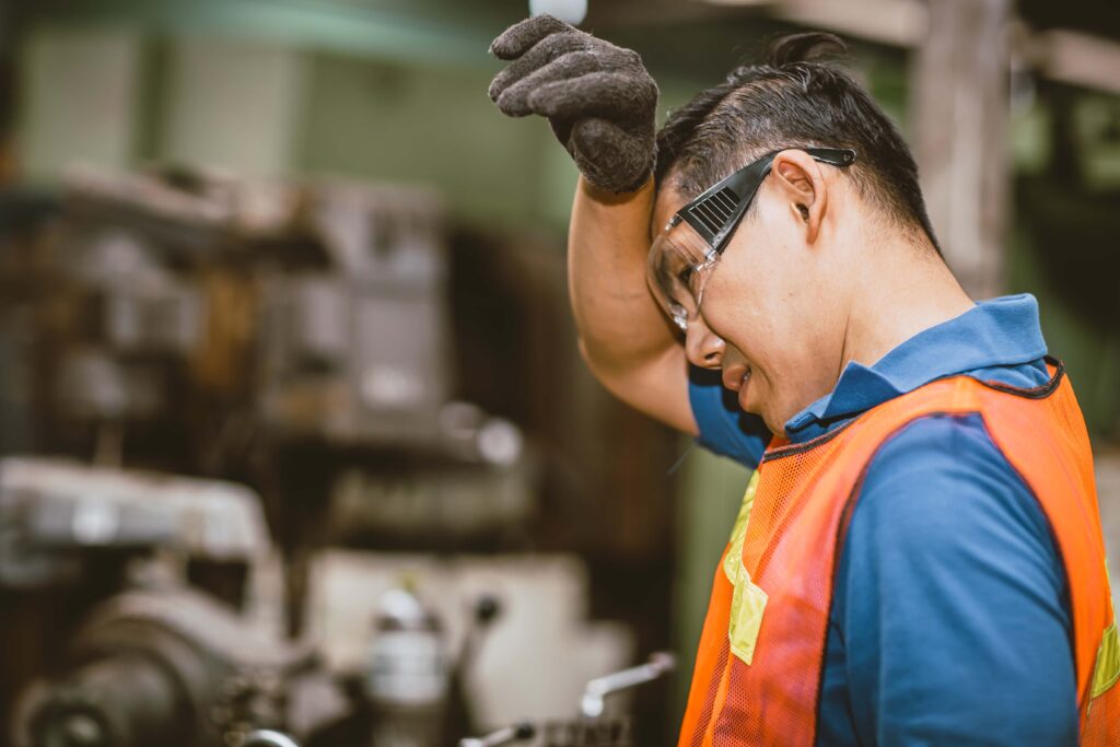 A male factory worker wearing safety goggles, a blue shirt, and an orange safety vest wipes his brow with a gloved hand. He appears tired and is standing inside an industrial setting with machinery in the background—an indication to spot early warning signs of heat stroke.