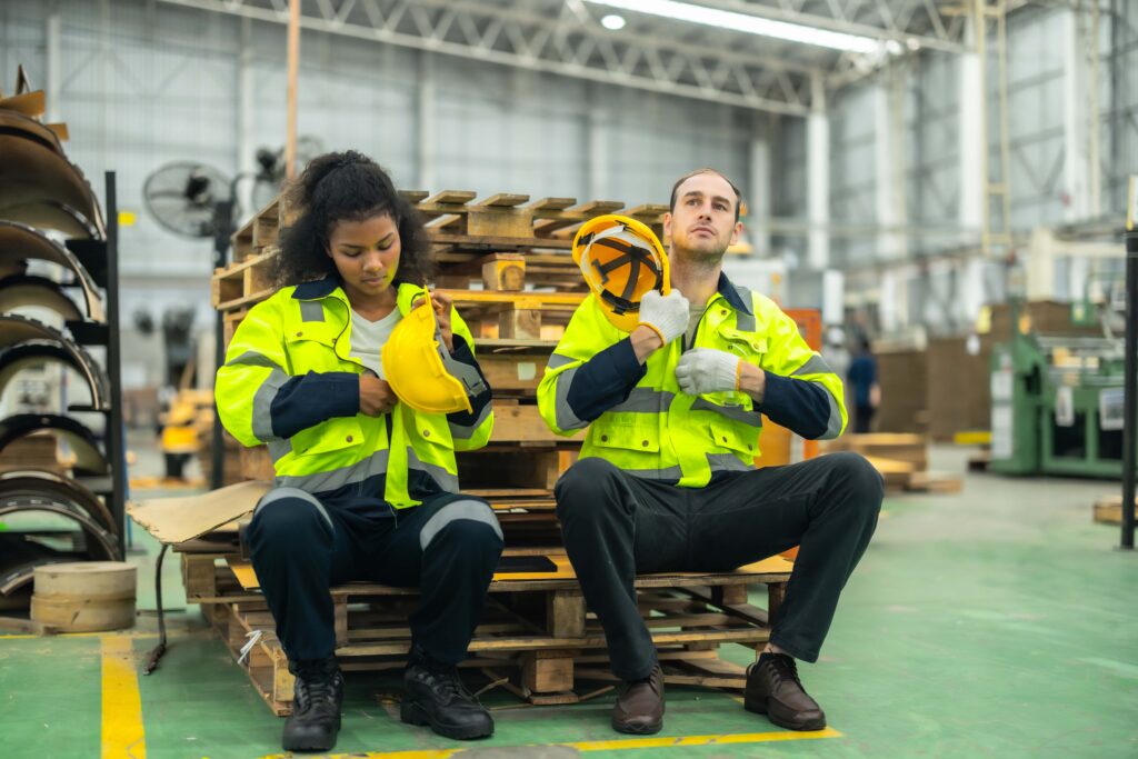 Two workers in high-visibility jackets are sitting on wooden pallets in an industrial warehouse. One is securing a hard hat, and the other is adjusting gloves, reflecting their commitment to PPE solutions. They appear to be preparing for work or taking a break in a busy, well-lit workspace.