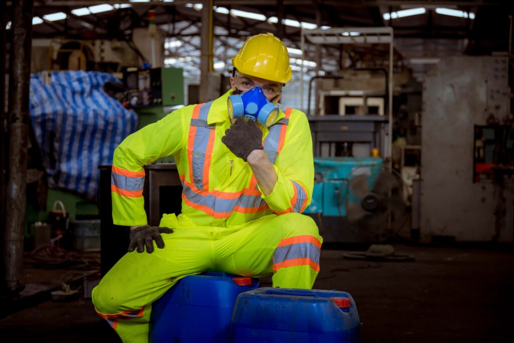 A worker in a neon yellow safety suit with reflective stripes, hard hat, and respirator mask sits on blue barrels in an industrial setting. Emphasizing the proper use of PPE, the worker holds the mask to their face. Machinery and equipment are visible in the background, highlighting this high-risk environment.