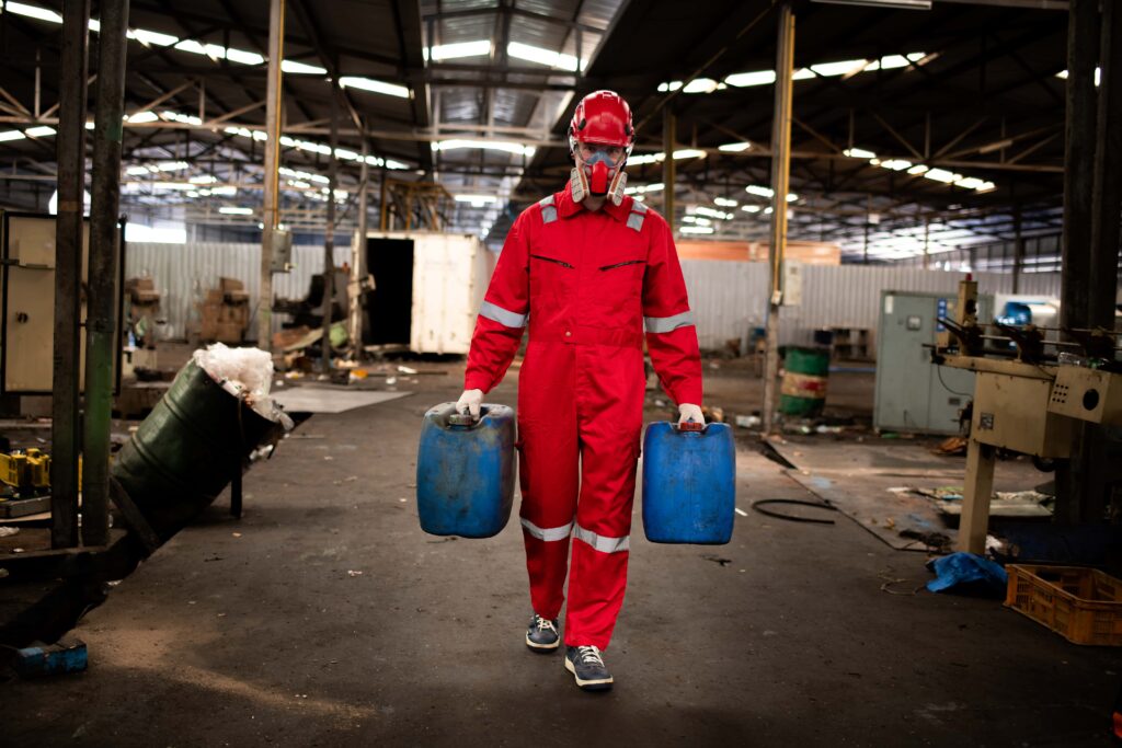 A person in a red protective suit, helmet, and face mask, carrying two blue containers walks through an industrial warehouse with various equipment and scattered items in the background. Their full-body protection ensures safety amidst the bustling environment.