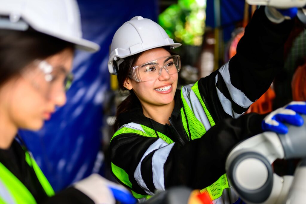 Two workers wearing safety helmets, face protection, and reflective vests operate machinery in an industrial setting. One worker in the foreground appears focused on a task while the other in the background smiles and adjusts equipment.