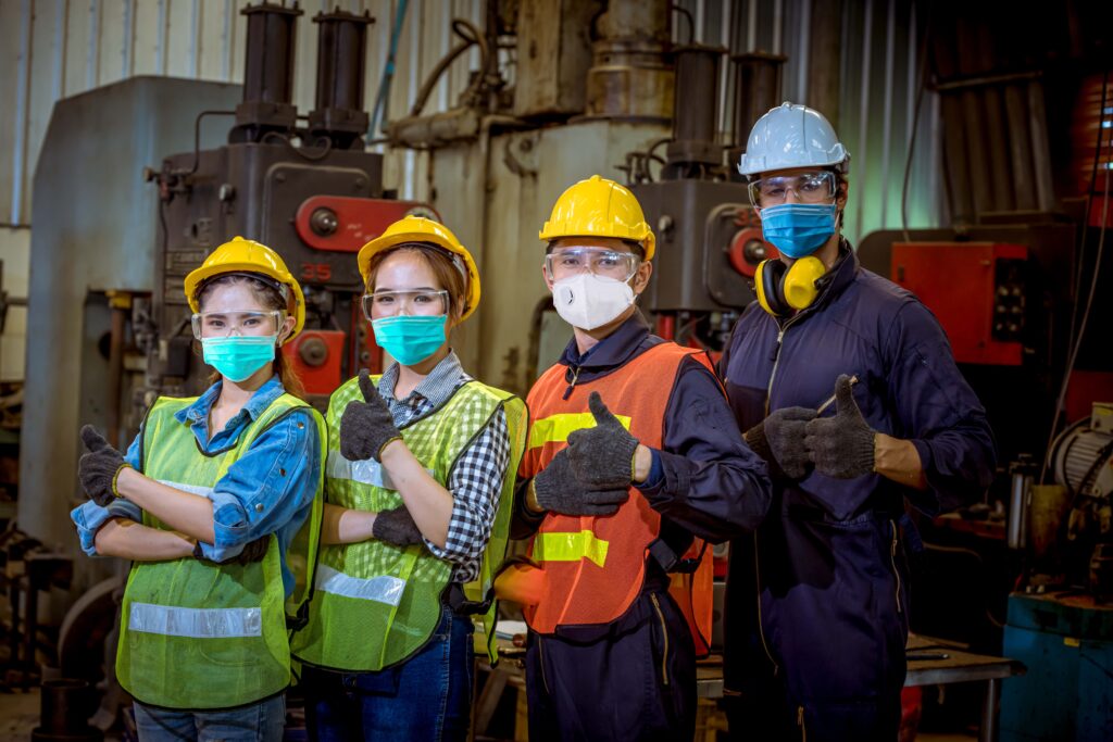 Four industrial workers wearing safety helmets, vests, gloves, and face masks pose with thumbs up in a factory setting. Heavy machinery and metal structures are visible in the background, exemplifying workplace safety and compliance with OSHA PPE standards.