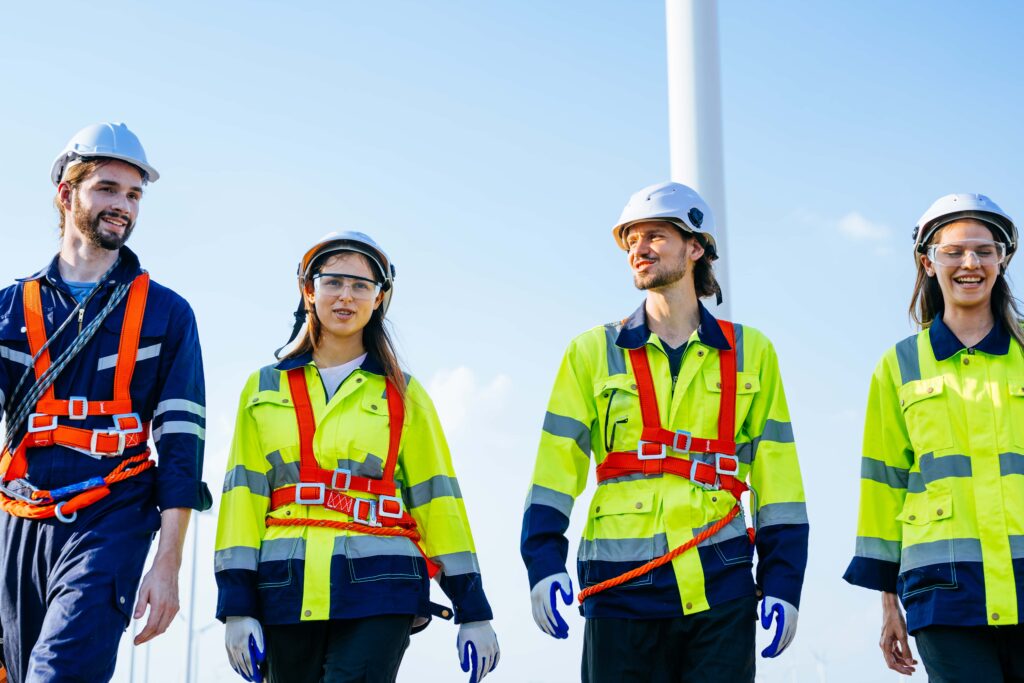 Four construction workers, suiting up safely with hard hats, high-visibility jackets, and safety harnesses, stand together outdoors under a clear blue sky. They appear to be walking and conversing about best practices, with wind turbines visible in the background.