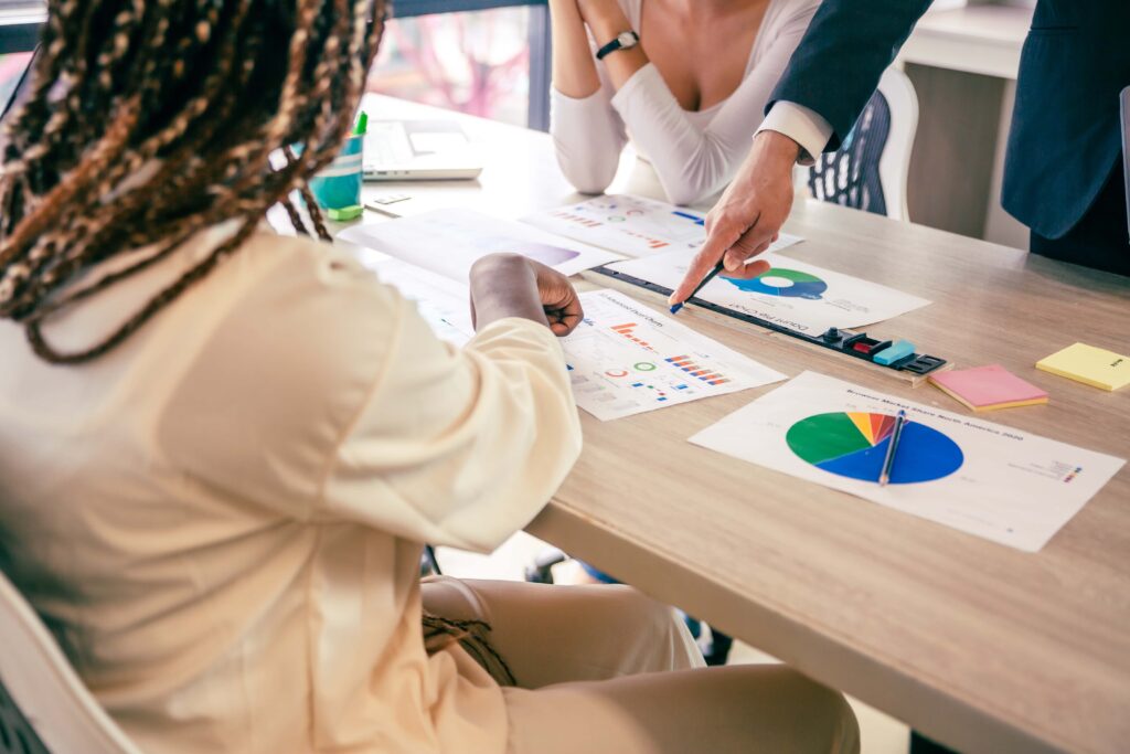 Three people sitting around a table are reviewing printed charts and statistics. One person is pointing at a document with a pen, while another is taking notes. The table is covered with various papers and office supplies, highlighting data on the improper use of PPE.