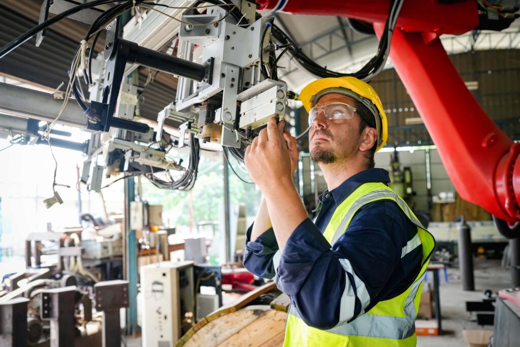 A male worker wearing a yellow hard hat and a high-visibility vest inspects machinery in an industrial setting. Ensuring proper PPE use, he is adjusting components connected to a robotic arm or automated system in a brightly lit workshop or factory.