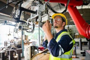 A male worker wearing a yellow hard hat and a high-visibility vest inspects machinery in an industrial setting. Ensuring proper PPE use, he is adjusting components connected to a robotic arm or automated system in a brightly lit workshop or factory.