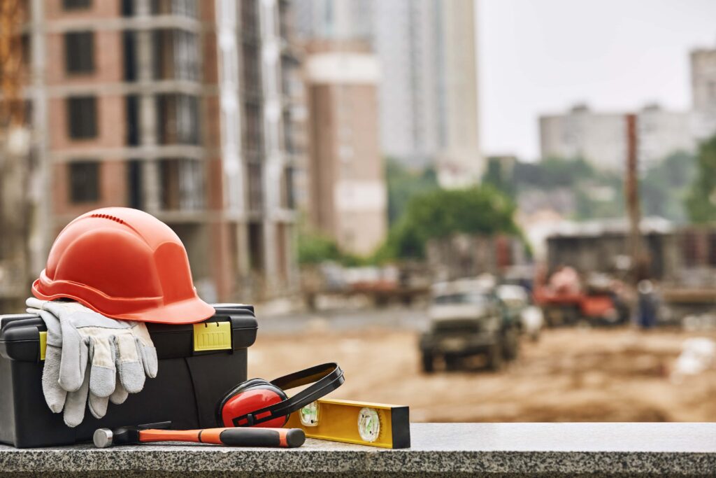 A toolbox with safety gear, including a hard hat, gloves, ear protection, and a spirit level, sits on a ledge at a construction site. In the background, there's blurred construction activity with buildings, machinery, and workers.