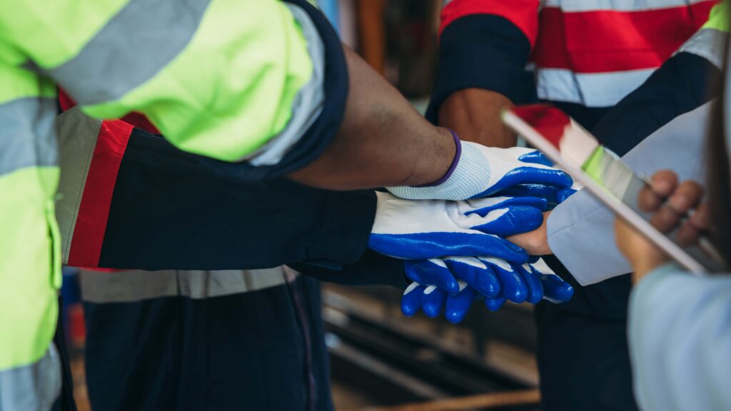 A close-up of several workers in a dynamic workplace, wearing high-visibility clothing and PPE gloves, stacking their hands together in a show of teamwork. One person holds a tablet, indicating a collaborative work environment.