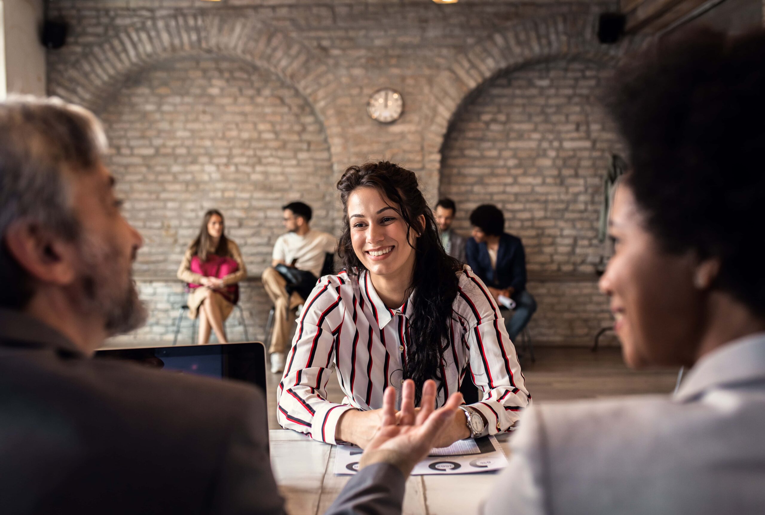In a brick-walled room with large archways, a person with long dark hair sits at a table smiling and discussing workplace health with two colleagues. In the background, other individuals sit and interact on benches, enjoying a shared sense of wellness. A clock is visible on the wall.