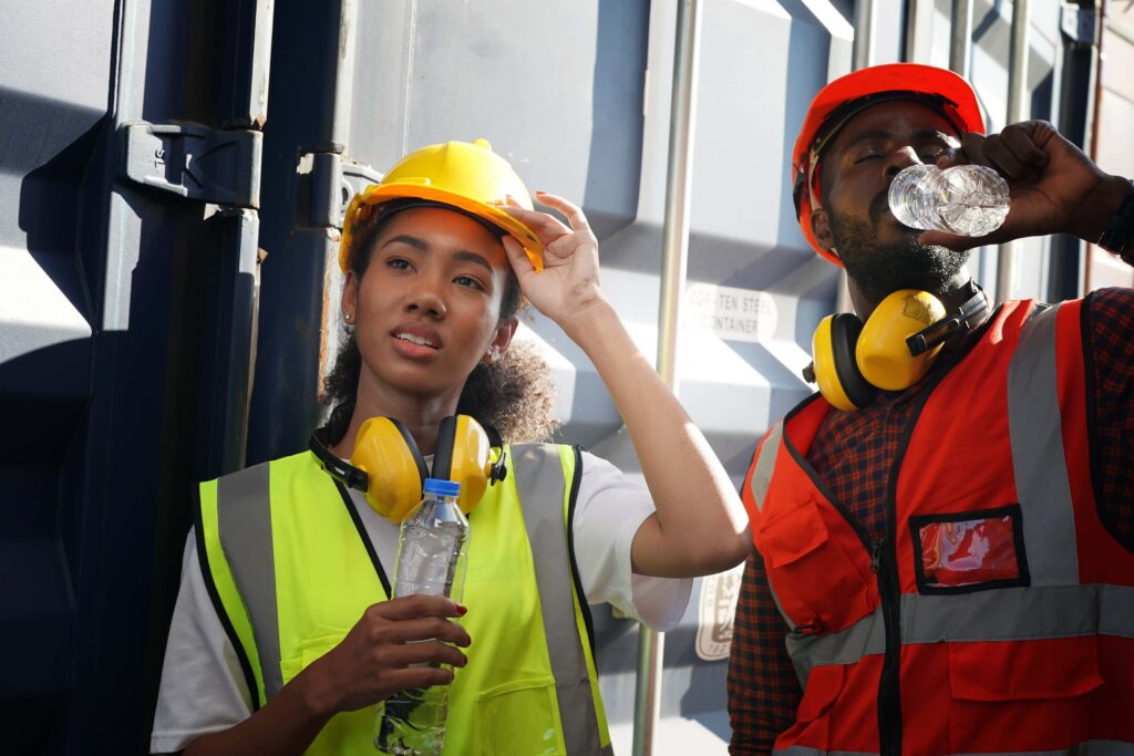 Two construction workers taking a break. One person, wearing a yellow safety helmet and vest, is holding a water bottle and adjusting their helmet. The other, in a red helmet and vest, is drinking water from a bottle. Both have ear protection around their necks. Humidity sensors play a crucial role in ensuring job site safety.