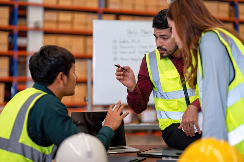 Three warehouse workers wearing safety vests and helmets engage in a discussion around a table with laptops. One man points with a pen while talking about risk reduction, another listens attentively, and a woman stands leaning towards them. Shelves and a whiteboard are visible in the background.