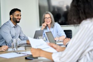 A diverse group of four colleagues are sitting around a conference table during a meeting. Two people, a man and an older woman with glasses, are facing the camera, smiling, and engaging in conversation about employee mental health. Laptops and key statistics on documents are on the table.