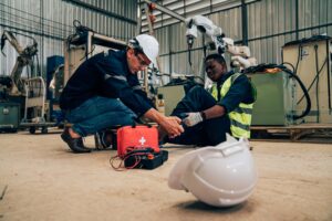 A construction worker in a white helmet is kneeling and providing first aid to an injured colleague, who is sitting on the floor holding his leg, inside an industrial facility. A first aid kit and a white helmet are on the ground beside them.