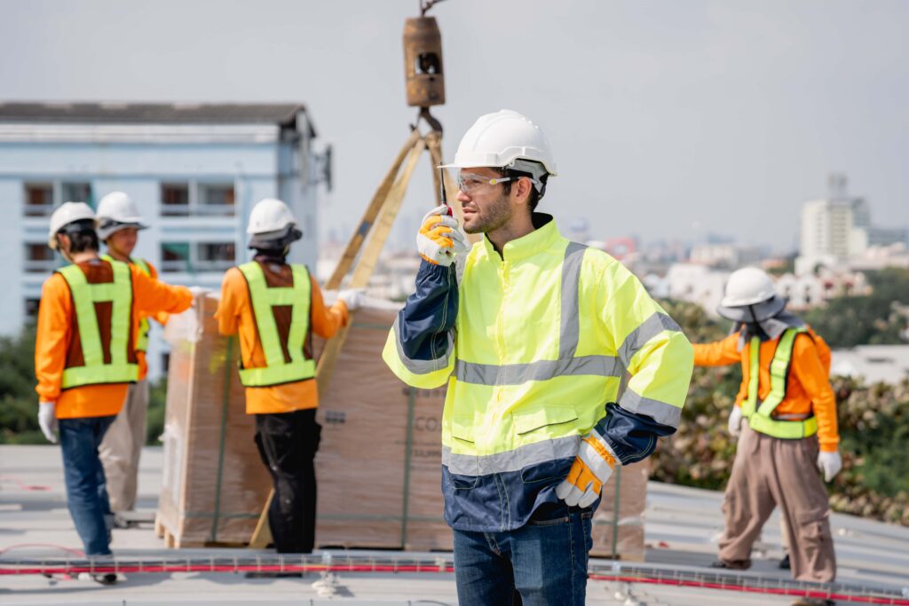 A construction worker in a yellow safety jacket, white hard hat, and protective glasses uses a walkie-talkie on a construction site. Promoting a culture of wellness and safety, three other workers in protective gear handle materials with a crane. Buildings are visible in the distance.