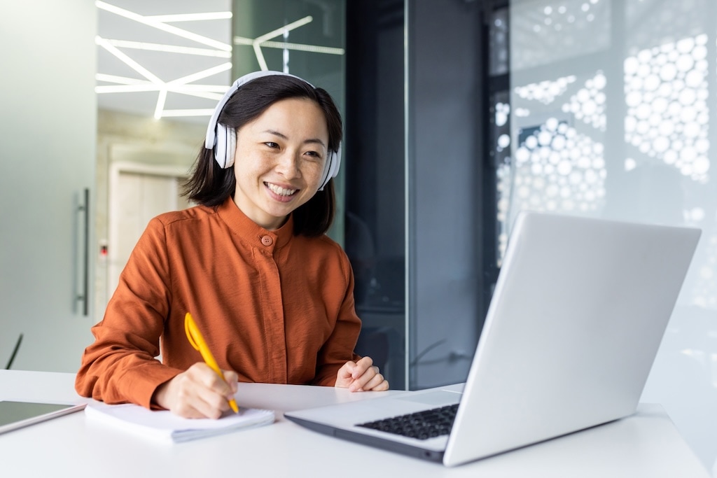 A woman wearing white headphones and a rust-colored blouse smiles while sitting at a desk. She jots down notes with a yellow pencil in front of her laptop, perhaps drafting ideas for an auto project. A modern geometric light fixture adds a touch of style in the background.