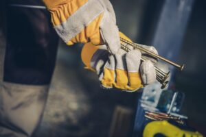 A person wearing yellow work gloves holds several large screws, keeping their hands warm while working outdoors. A construction area is visible in the background, with various tools and materials around, suggesting active work or preparation for a project.