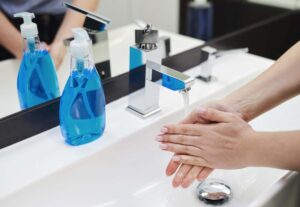 A person practices hand safety by washing their hands under a modern sink faucet. On the countertop, there is a bottle of blue liquid soap. The sink area is clean and well-lit, with a mirror reflecting part of the scene, ensuring optimal hygiene and skin care.