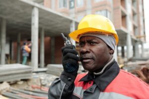 A construction worker, bundled up for the cold weather in a yellow hard hat and warm clothing, communicates via walkie-talkie at a busy site. Amidst work risks, a partly constructed building and another worker appear in the background.