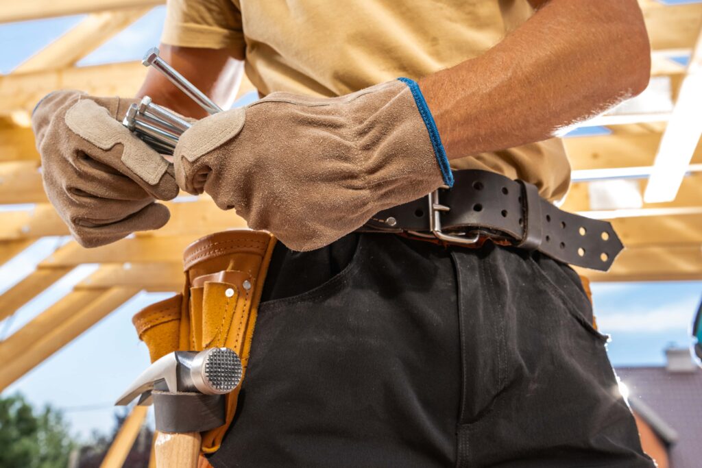 A construction worker wearing gloves and a tool belt with a hammer and other tools adjusts metal pipes. The worker stands against a background of partially constructed wooden framing under a clear, sunny sky.