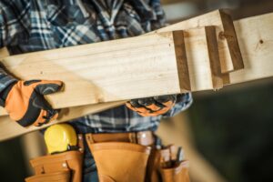 A person wearing a plaid shirt, gloves for hand protection, and a tool belt with various tools holds several wooden planks. The background is blurred, focusing on the individual and the wooden materials.