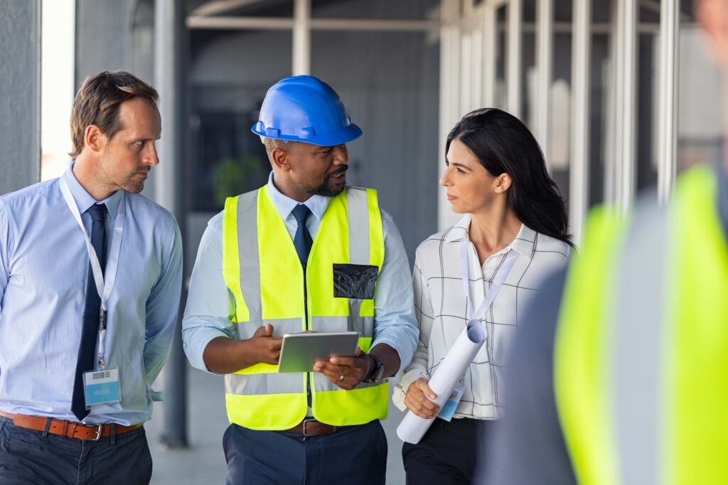 Three professionals at a construction site are discussing plans, emphasizing ThreeSixtySafety. A man in a blue helmet and high-visibility vest holds a tablet, while a woman in a white blouse holds rolled-up blueprints. Another man, also in professional attire, listens attentively.